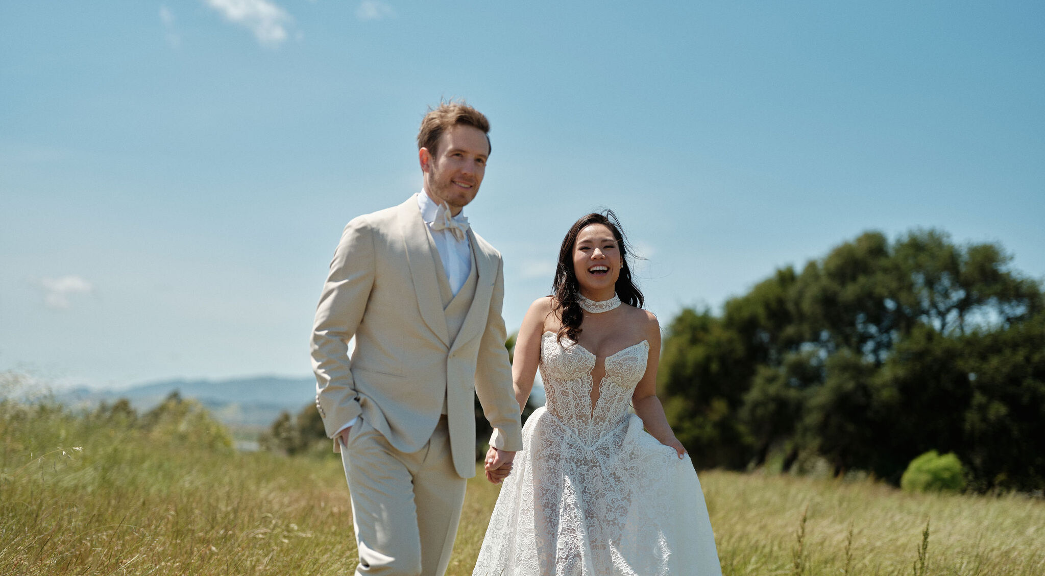 Bride Heidi with voluminous, flowing Kohnur romantic wedding hairstyle and radiant soft glam bridal beauty makeup, walking with groom Brandon in a sunny field