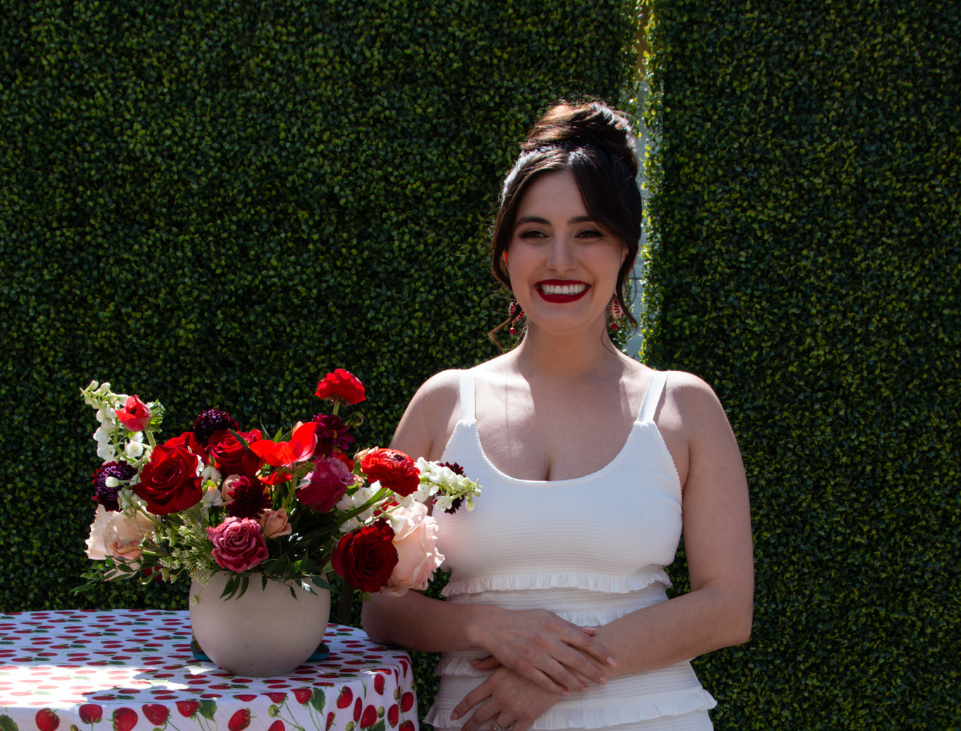 Roxanne with a chic, high-volume Kohnur elegant bridal updo and bold, vibrant soft glam bridal beauty makeup (red lip), posing next to red and white roses.